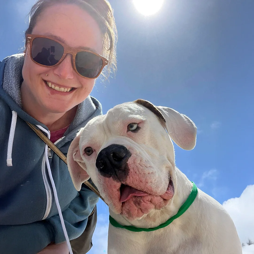 Person wearing sunglasses smiling beside a large white dog with a green collar, both posing outdoors under a sunny blue sky.
